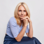 woman-with-blonde-hair-keeps-hands-near-face-looks-directly-camera-wears-blue-jumper-skirt-sits-white-thinks-about-something
